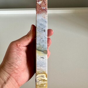Op de afbeelding: Een hoge, rechthoekige stenen obelisk met een puntige top. De steen heeft een mix van kleuren, waaronder rood, wit, grijs en geel. De obelisk wordt in een hand gehouden, wat de grootte en vorm laat zien. Dit decoratieve item is geschikt voor woondecoratie.