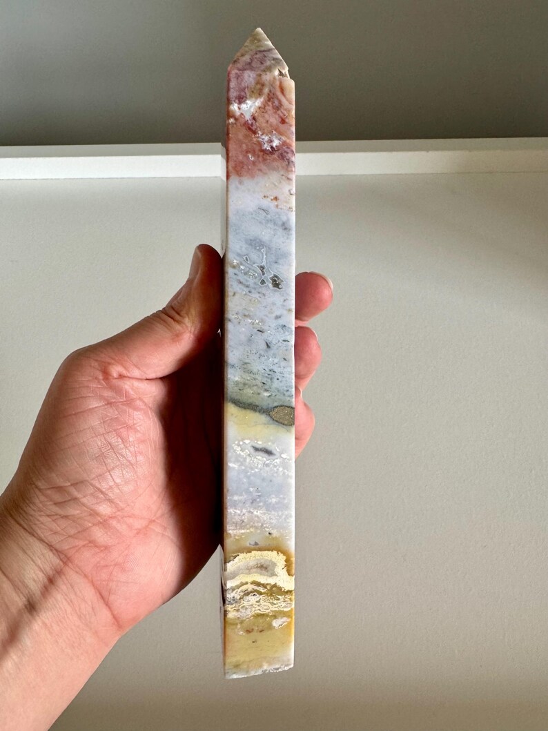 Op de afbeelding: Een hoge, rechthoekige stenen obelisk met een puntige top. De steen heeft een mix van kleuren, waaronder rood, wit, grijs en geel. De obelisk wordt in een hand gehouden, wat de grootte en vorm laat zien. Dit decoratieve item is geschikt voor woondecoratie.