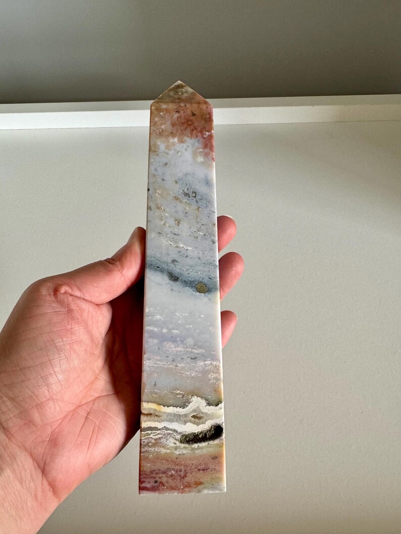 Op de afbeelding: Een rechthoekige kristallen obelisk met een puntige top. De steen heeft een mix van kleuren, waaronder wit, grijs, bruin en rood, waardoor een natuurlijk, gelaagd patroon ontstaat. De obelisk wordt in een hand gehouden, wat het gepolijste oppervlak benadrukt.