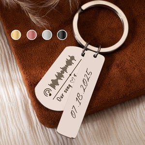 May include: A silver keyring with two rectangular tags. One tag features a soundwave design and the words "Our song ♡s". The other tag has the date "07.18.2025". The keyring is on a brown surface with a soft, textured background.