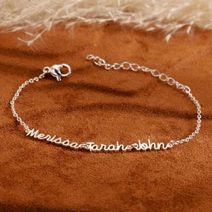 Op de afbeelding: Een delicate zilveren armband met de namen "Merissa", "Sarah" en "John" sierlijk over de ketting geschreven. De armband heeft een karabijnsluiting en een verstelbare ketting. De sieraden worden gepresenteerd op een bruin oppervlak met een zachte, neutrale achtergrond.