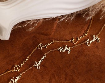 May include: Gold-colored name necklaces with the names "Kevin", "Dana", "Simon", "Piper", "Ruby", "Elvis", and "Emily" in a cursive font. The necklaces are displayed on a brown surface with a white decorative vase in the background.