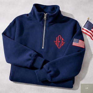 May include: Navy blue sweatshirt with a quarter-zip closure and a red embroidered monogram. The sweatshirt features an American flag patch on the sleeve and is made of a soft, comfortable material.