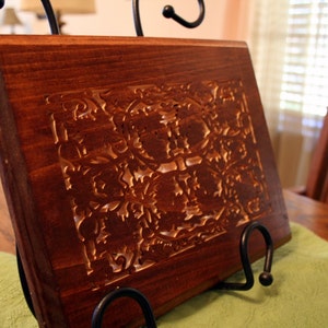 Op de afbeelding: Een donkerbruin houten snijplank met een ingewikkeld gesneden ontwerp. De plank ligt op een groene doek en een zwarte metalen standaard.