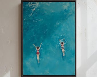 May include: A framed rectangular wall art print depicting an aerial view of two individuals swimming in vibrant turquoise water. One swimmer wears a red two-piece swimsuit, while the other wears a patterned two-piece swimsuit, both with arms raised.