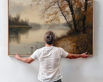 May include: A person stands in front of a framed landscape painting. The painting depicts a river scene with trees and a path leading to the water. The sky is a muted gray with hints of blue. The painting is framed in a light wood frame.