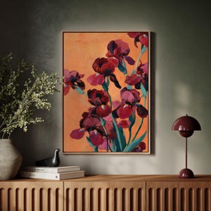 May include: An abstract painting of burgundy irises against an orange background. The flowers have green stems and leaves. The artwork is in a gold frame and hangs on a dark green wall. A burgundy lamp sits on a wooden cabinet.