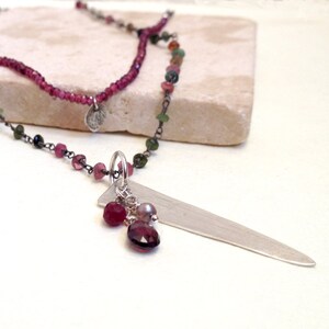 May include: Two layered necklaces with a silver pendant and gemstone beads. The top necklace is a strand of small, deep pink beads. The bottom necklace has a silver pendant with a cluster of gemstones, including a large, deep red stone.