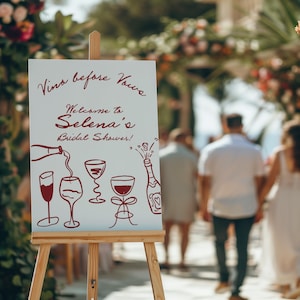 May include: A white sign on a wooden easel with the text "Vino before Vows" and "Welcome to Selena's Bridal Shower!" in burgundy script. The sign features illustrations of wine glasses and a champagne bottle.