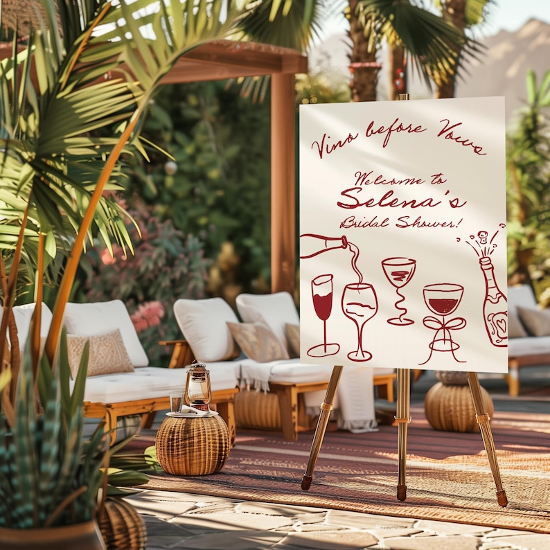 May include: A white sign on a wooden easel with the text "Vino before Vows" and "Welcome to Selena's Bridal Shower!" in burgundy script. The sign features illustrations of wine glasses and a bottle. The scene is set outdoors with lounge chairs.