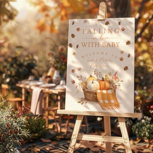 May include: A wooden easel displays a baby shower welcome sign. The sign features the text "Falling in love with baby" and "Welcome to Linda & Michael's Baby Shower." A basket of pumpkins and a plaid blanket are illustrated below the text.