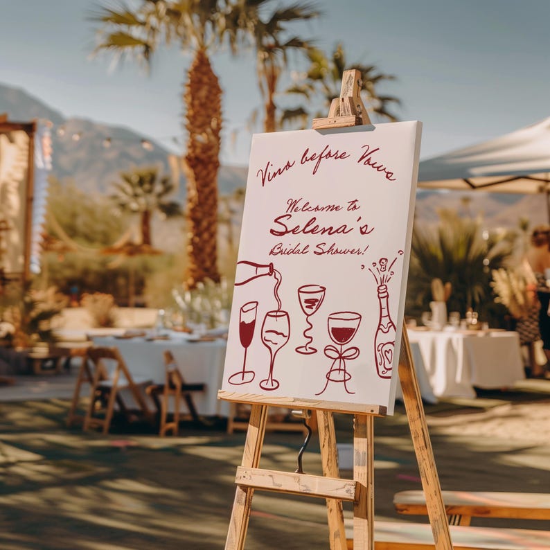 May include: A white sign on a wooden easel with the text "Vino before Vows" and "Welcome to Selena's Bridal Shower!" in burgundy script. Wine glasses and a bottle are illustrated below the text. The scene is outdoors with palm trees.