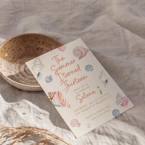 Pu&ograve; includere: Un invito color crema con illustrazioni di conchiglie e il testo "The Summer I Turned Thirteen" in rosa. L'invito &egrave; per la festa di compleanno di Selena, con data, ora e luogo.