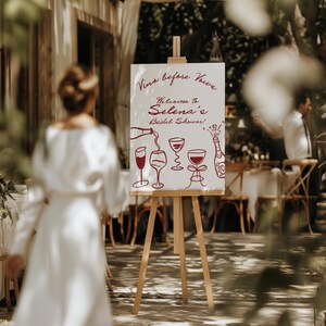 May include: A white sign on a wooden easel with the text "Vins before vows" and "Welcome to Selena's Bridal Shower!" in red script. The sign features red illustrations of wine glasses and a bottle.