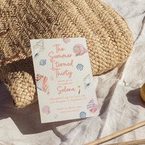 Puede incluir: Una bolsa de paja tejida beige descansa sobre una tela blanca junto a una invitaci&oacute;n de fiesta. La invitaci&oacute;n presenta ilustraciones de conchas marinas en acuarela y el texto "The Summer I turned Thirty" con detalles del evento.