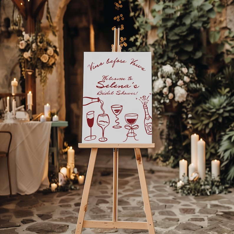 May include: A wooden easel displays a white sign with the text "Vino before Vows" and "Welcome to Selena's Bridal Shower!" in burgundy script. Wine glasses and a bottle are illustrated below. The sign is set in a garden setting.