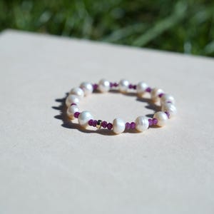May include: A white pearl bracelet with small purple beads and gold accents. The bracelet is circular and sits on a light beige surface. The pearls are irregularly shaped and have a natural luster.