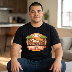 May include: A black t-shirt featuring a graphic of a brown sloth resting in a hammock between two trees. Above the sloth is a thought bubble with a low battery icon labelled 2%. Below the graphic, the text reads: CURRENTLY ON 2%—DO NOT DISTURB.
