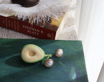 May include: Two pearl earrings with gold settings on a green surface next to a felt avocado.