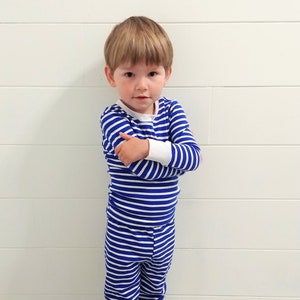 以下が含まれることがあります： 白と青のストライプのパジャマを着た幼い子供が、白い壁と白い腰板の前に立っています。子供は腕を組んでカメラを見ています。