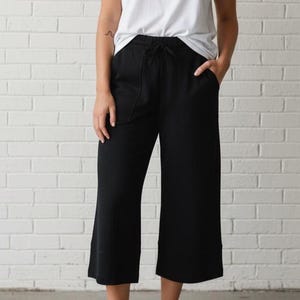 May include: A person wearing a white t-shirt and black culottes with pockets. The outfit is completed with white sneakers. The background is a white brick wall.