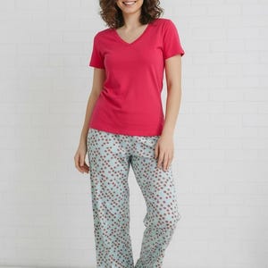 May include: A woman wearing a magenta V-neck t-shirt and light blue pajama pants. The pajama pants have a repeating pattern of small red dots. The background is a white brick wall.