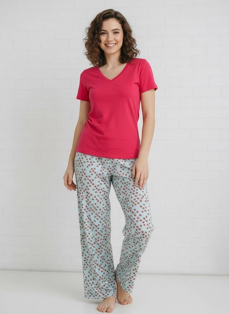 May include: A woman wearing a magenta V-neck t-shirt and light blue pajama pants. The pajama pants have a repeating pattern of small red dots. The background is a white brick wall.