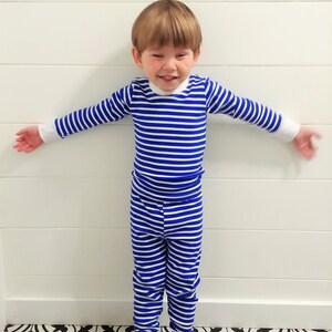 以下が含まれることがあります： 青と白のストライプのパジャマを着た幼い子供。子供は白黒のタイル張りの床の上に立っています。