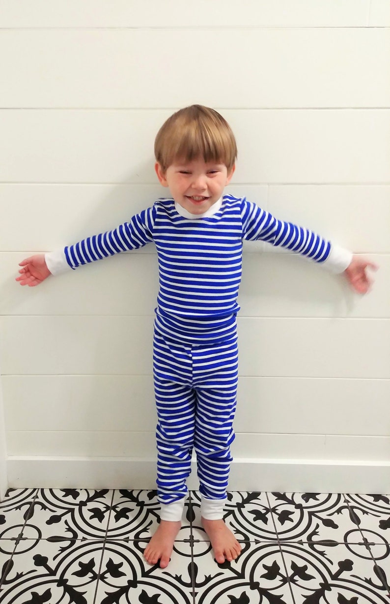 以下が含まれることがあります： 青と白のストライプのパジャマを着た幼い子供。子供は白黒のタイル張りの床の上に立っています。