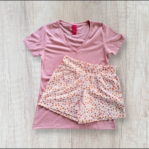 May include: A pink v-neck t-shirt with a floral print short. The shorts have a white background with small red, orange, and yellow flowers.