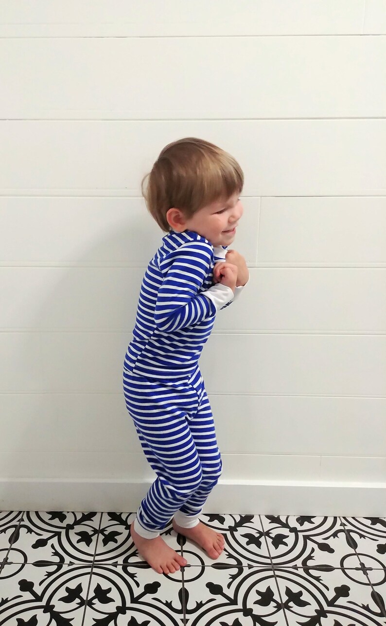 以下が含まれることがあります： 青と白のストライプの長いパジャマを着た小さな子供。子供は白黒の模様のタイル張りの床の上に立っています。