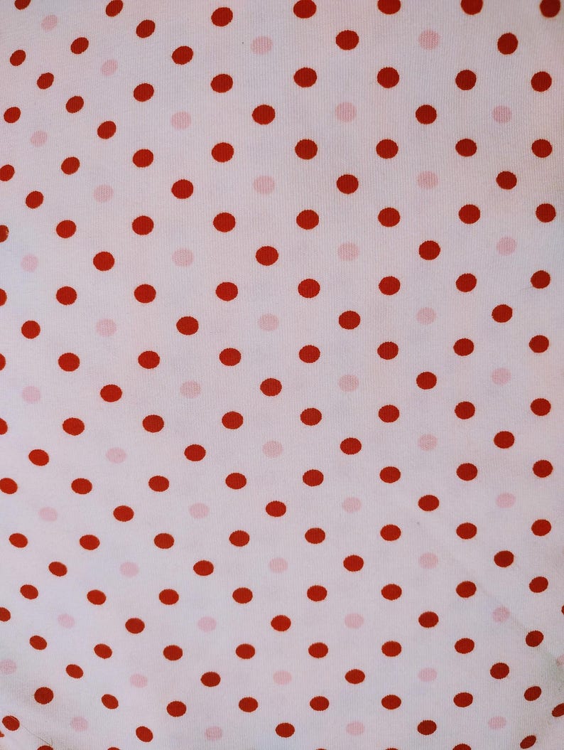 May include: Close-up of a white fabric with a repeating pattern of red polka dots. The dots are evenly spaced and vary slightly in size, creating a playful and classic design. The fabric appears to be soft and lightweight.