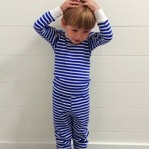 以下が含まれることがあります： 青と白のストライプのパジャマを着た小さな子供。パジャマは長袖で長ズボンです。子供は白黒のタイル張りの床の上に立っています。