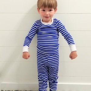 以下が含まれることがあります： 青と白のストライプのパジャマを着た小さな子供。パジャマは長袖で長ズボンです。子供は白黒のタイル張りの床の上に立っています。