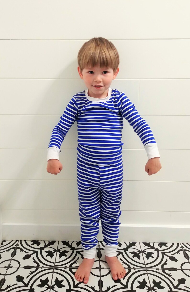 以下が含まれることがあります： 青と白のストライプのパジャマを着た小さな子供。パジャマは長袖で長ズボンです。子供は白黒のタイル張りの床の上に立っています。