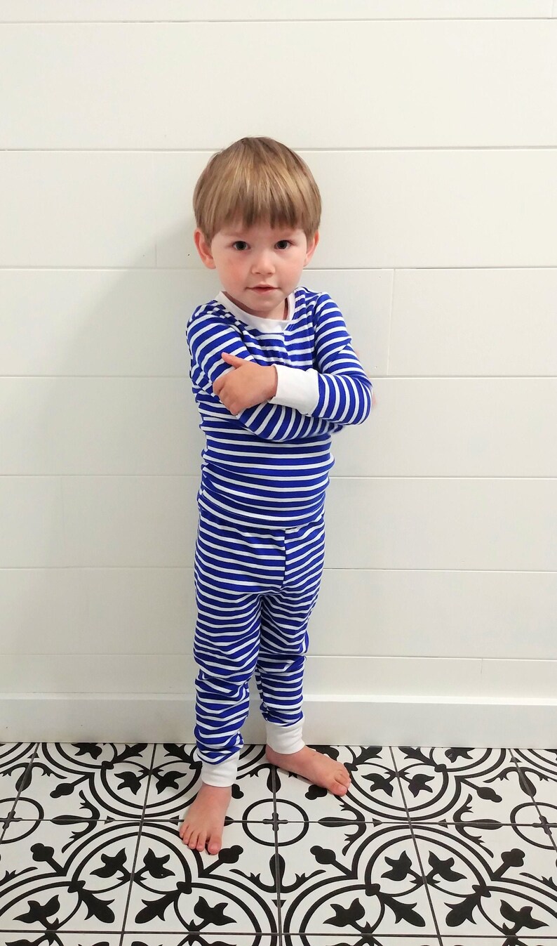 以下が含まれることがあります： 白と青のストライプのパジャマを着た幼い子供が、白い壁と白い腰板の前に立っています。子供は腕を組んでカメラを見ています。