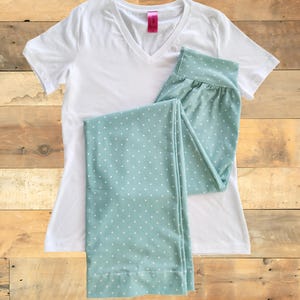 May include: A white V-neck t-shirt and light teal pajama pants with white polka dots are displayed on a wooden surface. The pajama pants are folded, and the t-shirt has a pink tag.