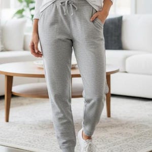 May include: Light gray sweatpants with a drawstring waist and pockets. The pants are paired with a white long-sleeve shirt and white sneakers. The outfit is casual and comfortable.