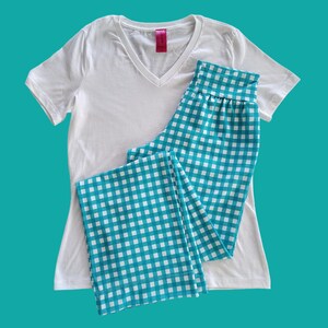 May include: A white V-neck t-shirt paired with turquoise and white checkered pajama pants. The pajama pants are folded and placed next to the t-shirt. The background is a solid turquoise color.