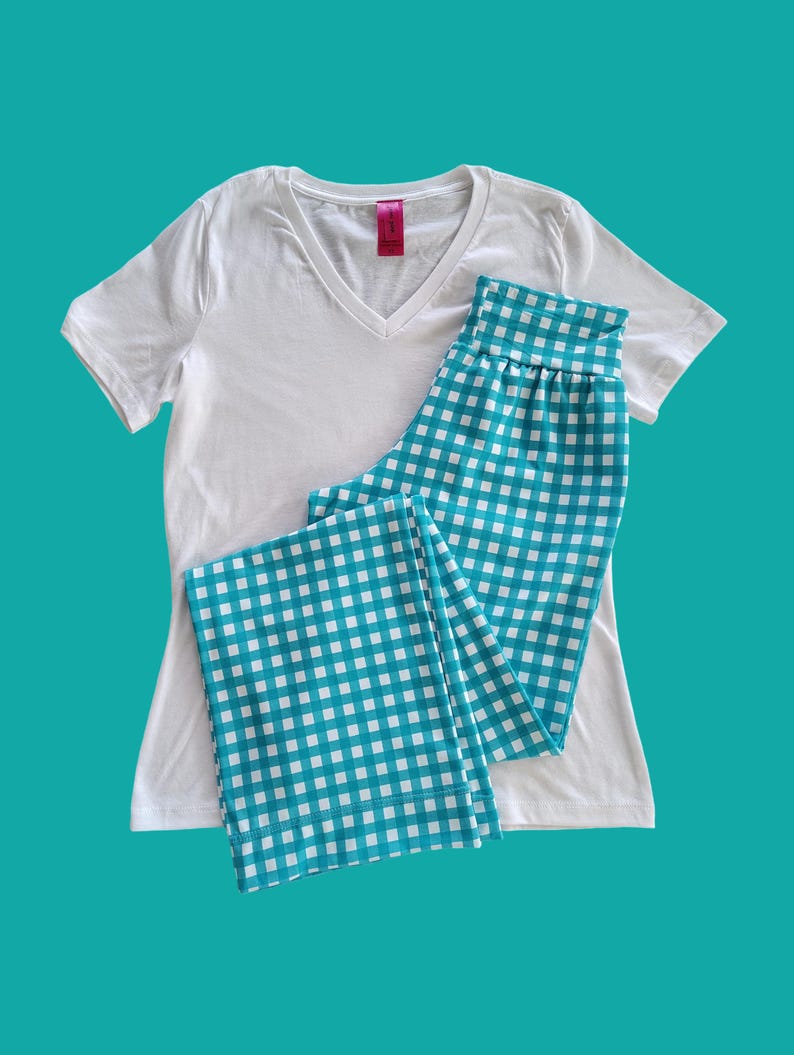 May include: A white V-neck t-shirt paired with turquoise and white checkered pajama pants. The pajama pants are folded and placed next to the t-shirt. The background is a solid turquoise color.
