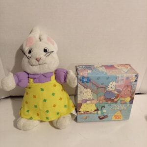 May include: A white plush rabbit toy wearing a purple shirt and a yellow dress with a green and orange clover pattern, positioned next to a square cardboard box featuring a 24-piece Max & Ruby puzzle with an illustration of the characters.