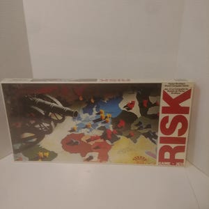 May include: A vintage box for the board game Risk. The box features a world map with colored territories and a cannon in the foreground. The text "RISK" is printed in large, bold letters on the box.