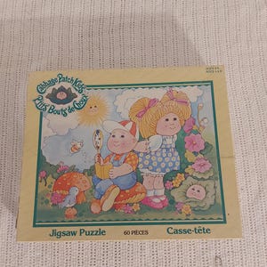 May include: A 60-piece jigsaw puzzle featuring Cabbage Patch Kids dolls in a garden setting. The puzzle box shows two dolls, one with blonde hair and a pink bow, and the other with brown hair and a blue shirt. The box also includes the text "Cabbage Patch Kids" and "Ptits Bouts de Choux".