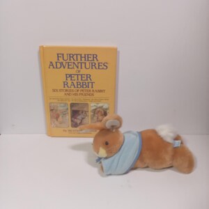 May include: A plush brown rabbit toy with a light blue neckerchief and a white tail, positioned in front of a yellow hardcover book titled Further Adventures of Peter Rabbit, Six Stories of Peter Rabbit and His Friends by Beatrix Potter.