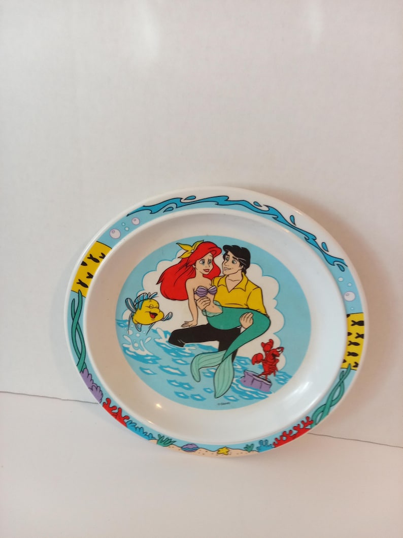 May include: A round white plate featuring a colorful illustration of a mermaid with red hair and a human character in a yellow shirt. The design includes a yellow fish, a red crab, and blue water. The rim is decorated with aquatic patterns.