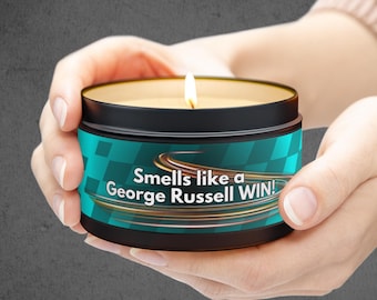 May include: A black candle with a teal and gold label that reads "Smells like a George Russell WIN!" The candle is lit and being held by a person's hands.