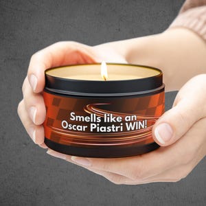 May include: A black candle with a red and orange label that says "Smells like an Oscar Piastri WIN!" The candle is lit and being held by a person's hands.