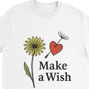 以下が含まれることがあります： 白いTシャツ。タンポポのデザイン。黄色の花、緑の茎、赤いハートと種が特徴です。「Make a Wish」の文字が黒でプリントされています。カジュアルな普段着のTシャツです。