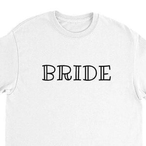 以下が含まれることがあります： 黒い縁取りの文字で「BRIDE」とプリントされた白いTシャツ。画像の下部にあるテキストは「Color White, Natural: Black Letter other Colors: White Letter」と書かれています。