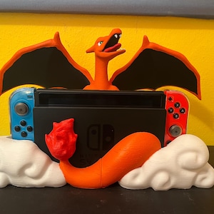 May include: A black Nintendo Switch console is flanked by blue and red controllers, with a 3D-printed orange Charizard figure and white cloud accents. The Charizard has black wings and is posed with its mouth open.